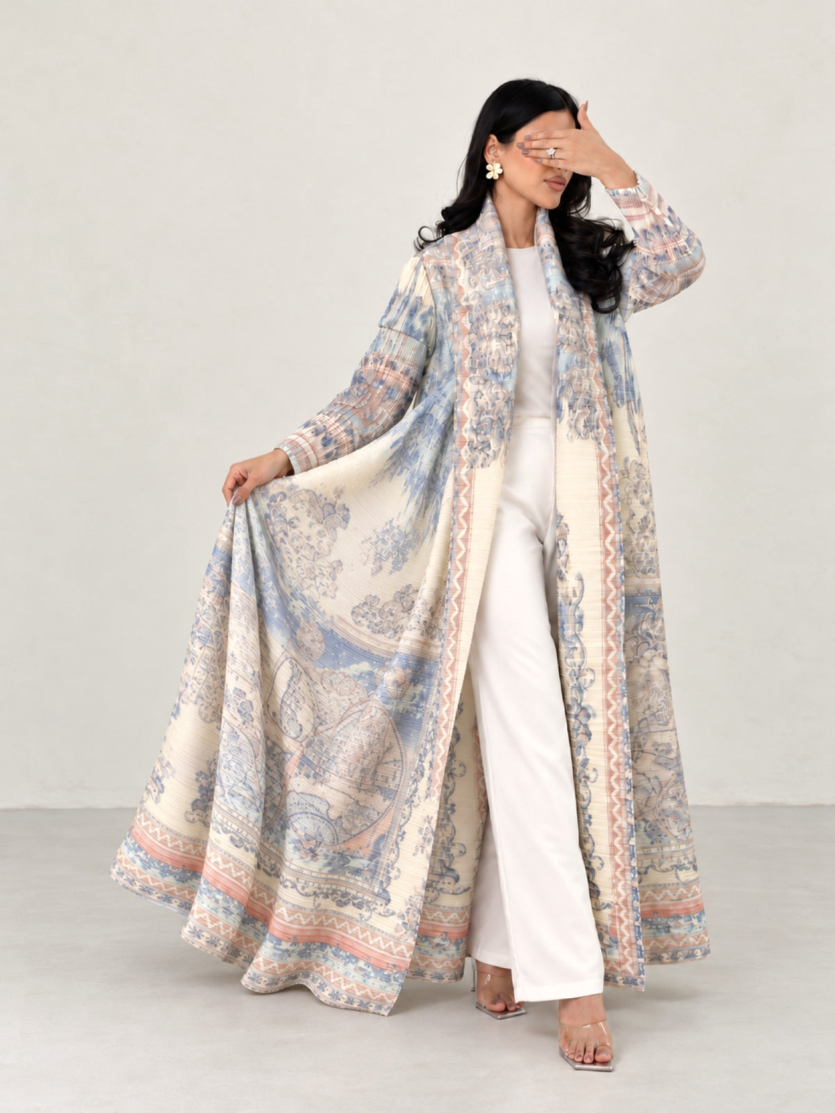 Model holding the Alamira printed open abaya out to the side, the fabric falling in soft folds, in an off-white studio